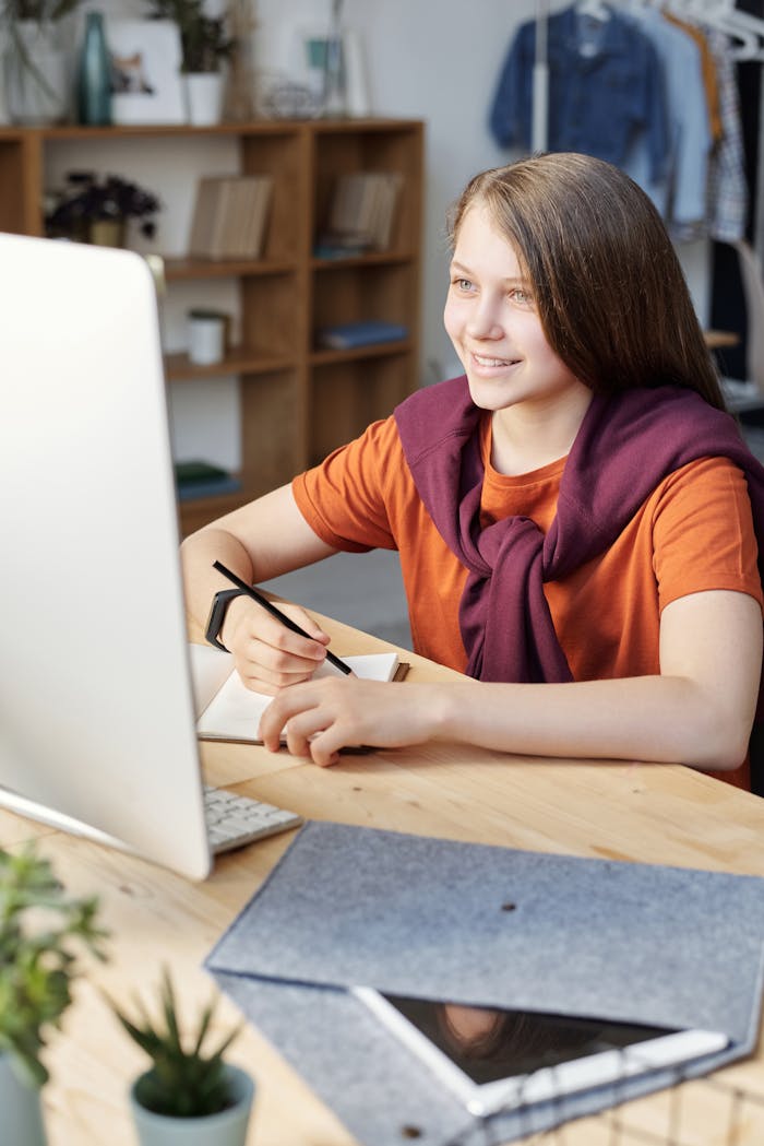 Smiling teen girl studying online at her home desk with a computer and notepad.